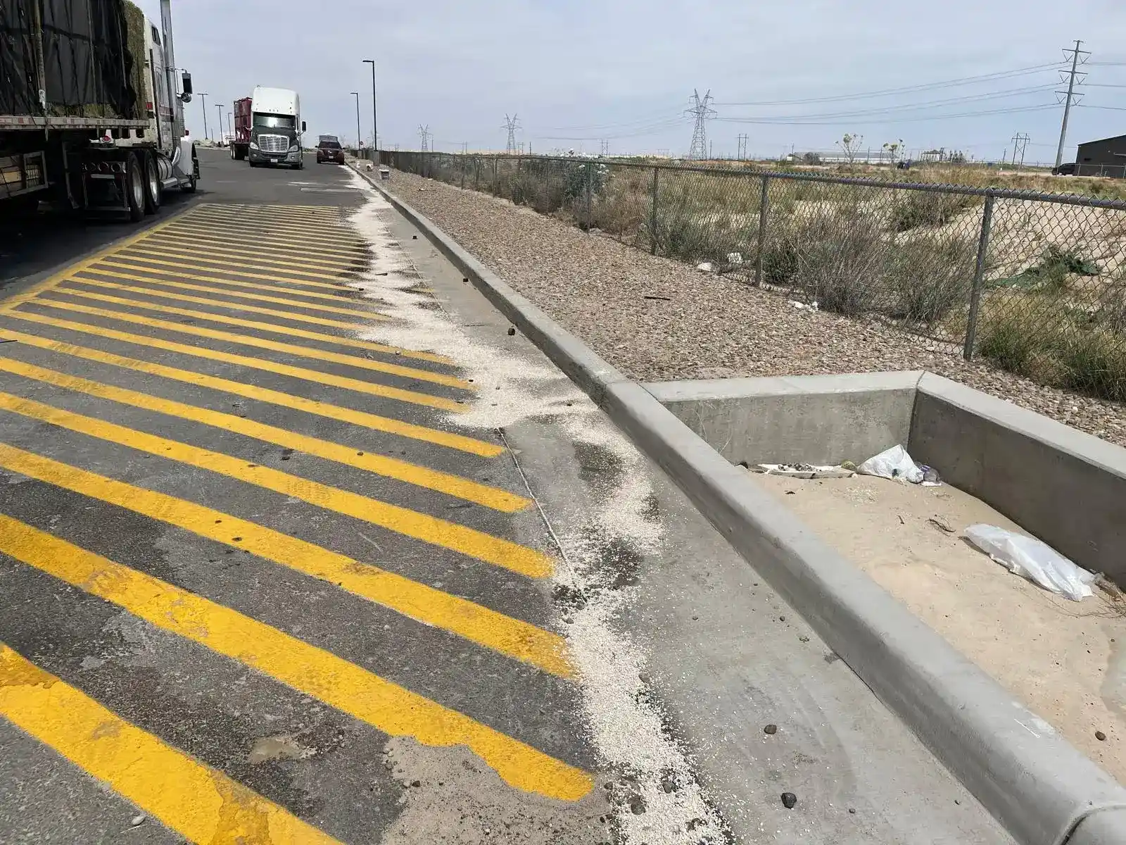 Industrial access road with yellow safety markings, trucks, and a concrete drainage basin